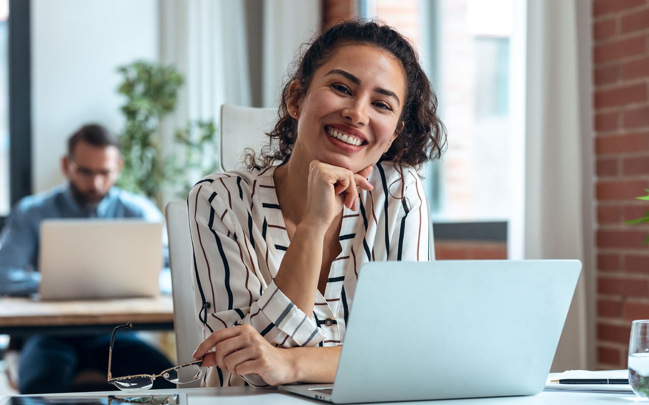 Smiling business woman working with laptop while looking at camera in modern startup office.